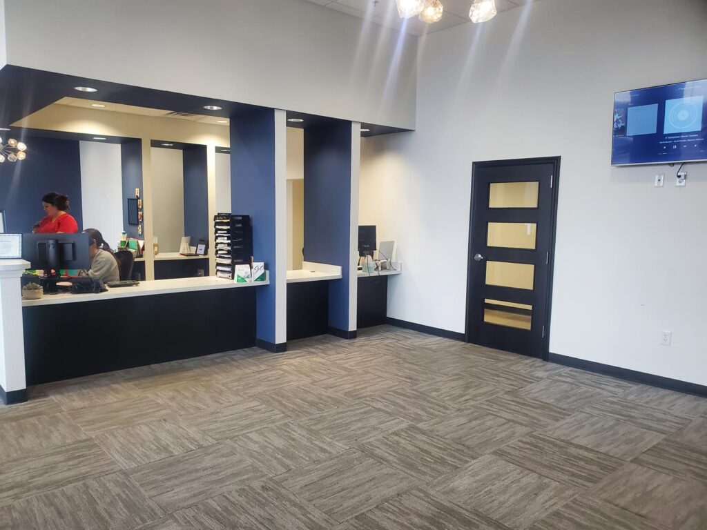 The image shows a reception area with a black and white counter, two workstations, and shelves for office supplies. The space features white and blue walls, grey carpet tiles, and recessed lighting for a clean, professional look.