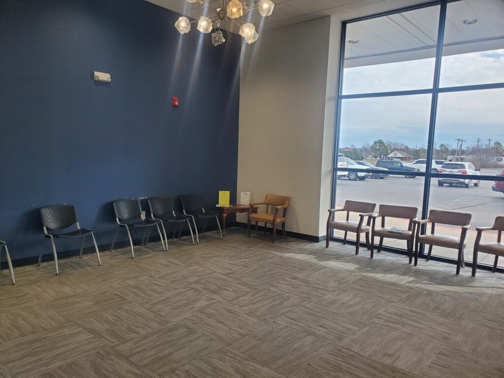 A waiting area with eight chairs arranged along the walls: four black chairs against a dark blue wall and four wooden chairs with armrests near a large window. A small table between the two sets holds items like a yellow sign and papers. The floor is carpeted, ceiling lights illuminate the space, and the view outside shows parked cars and nearby buildings.