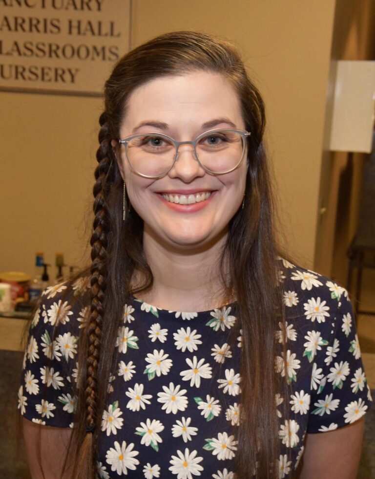A woman with long brown hair, partially braided on the left side, wearing a black shirt with a white daisy pattern. The background features a sign reading 'SANCTUARY HARRIS HALL CLASSROOMS NURSERY,' along with a table with some objects and a lamp.
