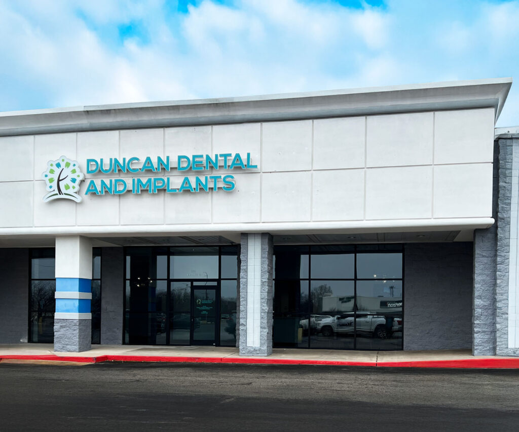 The image shows the exterior of a dental clinic named 'Duncan Dental and Implants.' The clinic features a modern design with large glass windows and a white and gray color scheme. Above the entrance, there's a prominently displayed teal and white sign with a logo featuring a stylized tooth and tree design. A red curb in front of the building indicates a no-parking zone.