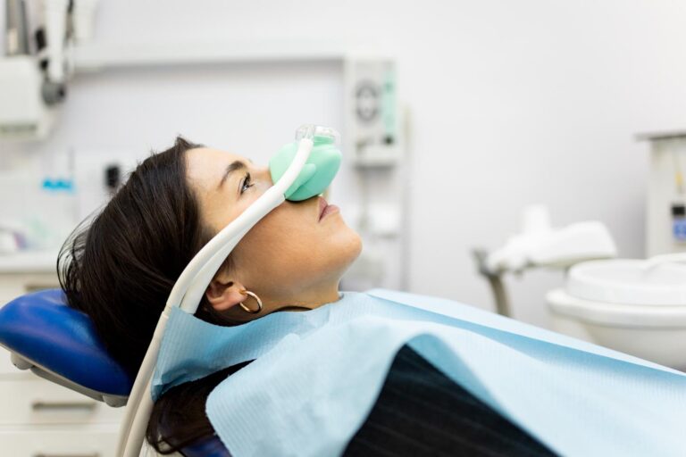 The image shows a person lying on a dental chair in a dental clinic. They are wearing a blue dental bib and appear to be undergoing a procedure or examination. The background features standard dental equipment, including a dental light, sink, and instruments, illustrating a typical dental clinic environment.