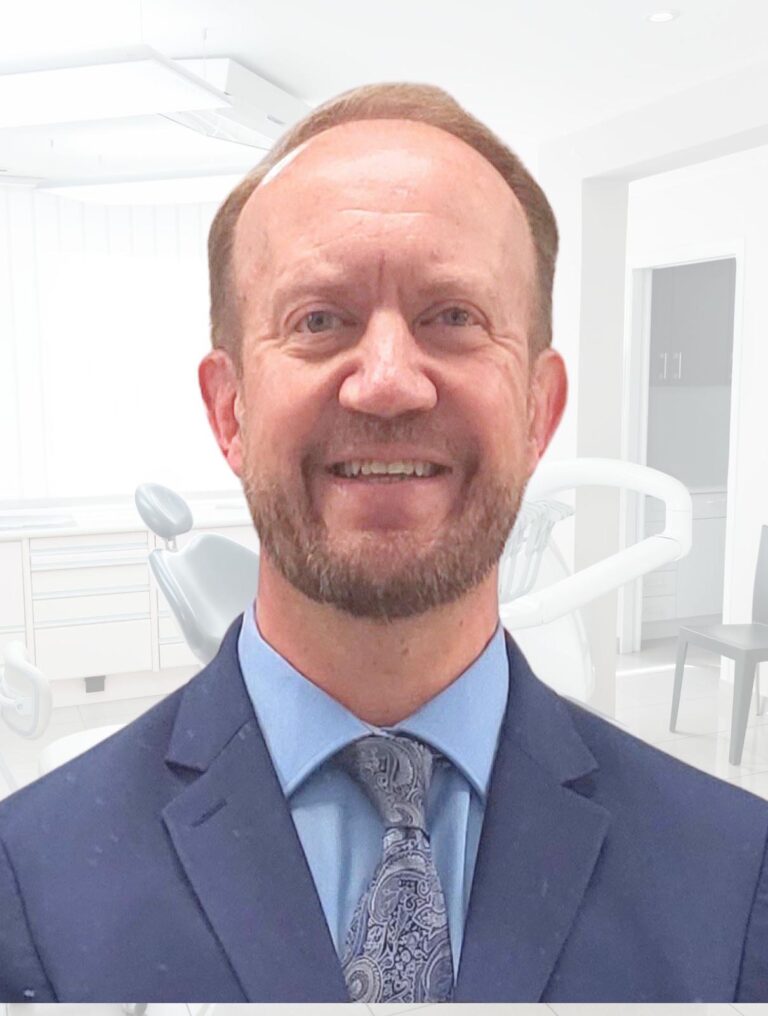 A man wearing a blue suit, light blue shirt, and patterned tie, standing in a dental office. The background includes dental equipment, cabinets, and chairs, creating a professional atmosphere.